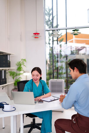 An Asian adult doctor in scrubs consults with a patient in a clinic using a laptop. Examination and treatment for illness. Healthcare professional helping patient in officeの写真素材
