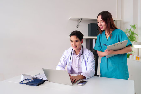An adult Asian doctor is using a laptop while an adult Asian nurse holds a chart. They are working together in a clinic office to discuss patient records and healthcare consultation.の写真素材