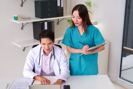 Asian adult doctor working on a laptop while an Asian young nurse stands by, reviewing notes on a clipboard. They collaborate on patient data for optimal healthcare outcomes. Teamwork in clinic office setting.の写真素材