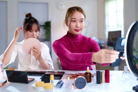Two young Asian women are creating beauty and cosmetics content for social media. They are streaming live, recording video for a makeup tutorial, and giving a product review in their home studio.の写真素材