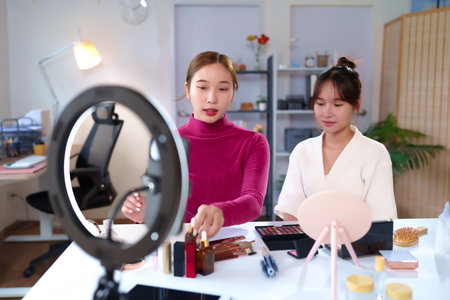 Two Asian young women presenting and demonstrating different beauty products and cosmetic on live streaming with ring light for social media online tutorial.の写真素材