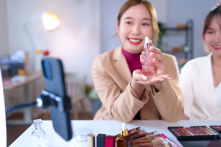 An Asian adult woman presents beauty serum product on live streaming. She is reviewing the skincare item with various makeup cosmetics nearby. Perfect for online beauty influencer content.の写真素材