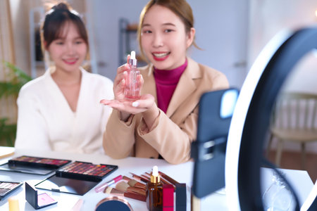 Two young Asian women demonstrate a skincare serum product during a live online beauty tutorial, sharing makeup application tips and promote cosmetic brand with ring light at home.の写真素材