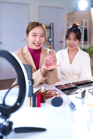 Two adult Asian women are broadcasting an online makeup tutorial. One presents a skincare product with a smile. Beauty products are on the table. Bright lighting helps present vlogの写真素材
