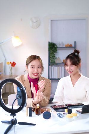 Two Asian women beauty bloggers are reviewing red lipstick and sharing cosmetic sales tips and recommendations online during a live stream for social media marketing in their studioの写真素材