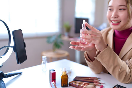 Adult Asian woman reviews a new beauty serum during her video stream for an online makeup tutorial to demonstrate new cosmetic products in a home studio setting for her fans.の写真素材