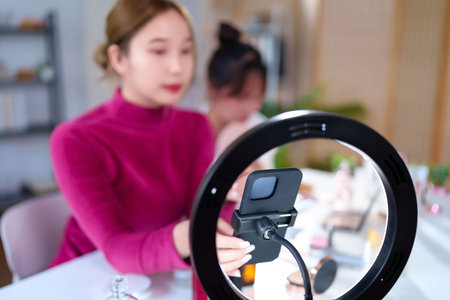 Two young Asian women are using a ring light and mobile phone to create beauty vlogging content for live streams and other social media platforms in her houseの写真素材