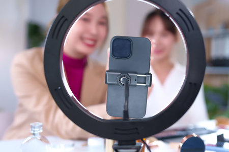 Two young Asian women are creating beauty content for social media. One is live streaming a makeup tutorial using cosmetics smartphone and ring light. Engaging content for beauty enthusiastsの写真素材