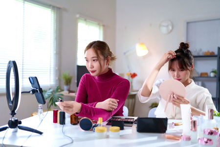 Two young Asian women creating beauty content for a vlog. One is applying makeup while streaming live tutorials with cosmetics. Focus on beauty and makeup for the audience online.の写真素材