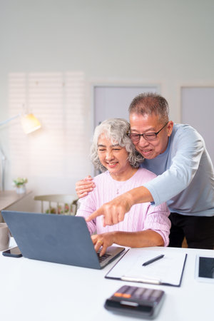 Cheerful older Asian couple using laptop together at home Happy senior couple looking at the screen browsing the internet shopping online technologyの写真素材