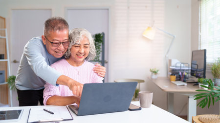 A loving older Asian couple is smiling at their laptop screen at home. They are enjoying their time together browsing online, filled with joy and happiness.の写真素材
