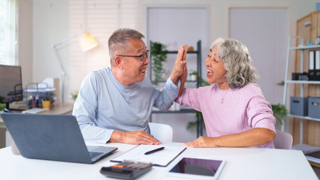Cheerful Asian older couple giving high five celebrating financial success at home. Enjoying retirement, planning savings, investments together with happiness and love.の写真素材
