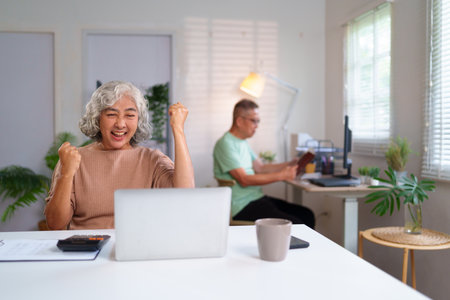 An older Asian woman joyfully celebrates a victory in her home office, using a laptop computer while her partner relaxes nearby in a shared retirement space.の写真素材