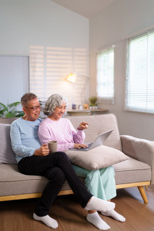 Older Asian couple enjoys quality time at home. They are relaxing on the sofa, using a laptop, smiling, and drinking coffee. Happy moments create lasting memories together.の写真素材