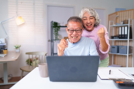Excited older Asian couple celebrates success at home. They are using a laptop on a desk with paperwork in their bright home office, sharing a moment of happiness.の写真素材