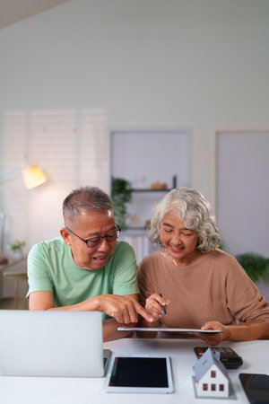 An older Asian couple happily reviews financial documents together at home using a laptop, tablet and calculator planning for future property investment and their retirementの写真素材