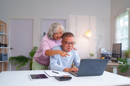Happy older Asian couple using a laptop at home. The wife points to the screen while the husband types. This charming moment shows retirement and connection.の写真素材