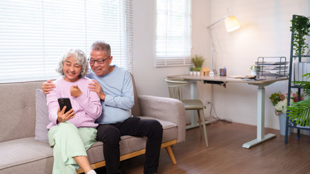 Adorable Asian older couple embracing their golden years with joy using a smartphone for entertainment and communication celebrating companionship and family love at homeの写真素材