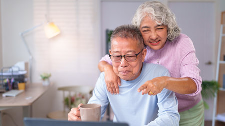 An older Asian couple uses a laptop together at their home. They share a moment of love and connection during retirement in their cozy living room.の写真素材