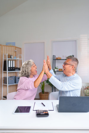 An older Asian couple expresses joy and achievement with a high five while working at their home office, creating a delightful retirement lifestyle moment.の写真素材