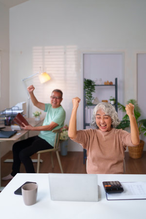 A happy older Asian couple celebrating a success at home with laptop and documents. They achieve goals together while managing their retirement funds.の写真素材