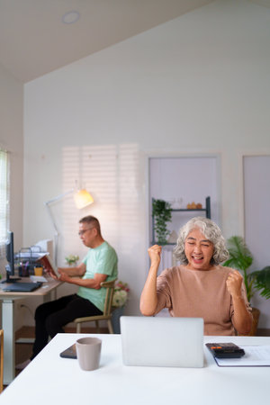 An older Asian woman is celebrating success in her home office while working on laptop, with a senior man reading a book in the background.の写真素材