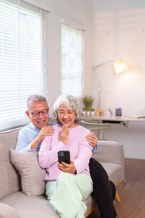 Asian older couple sitting together on sofa using smartphone for video call smiling at home Happy senior man and woman enjoying using mobile phoneの写真素材