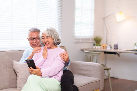 A delightful Asian older couple is happily engaged in a video call on their smartphone while sitting on a cozy sofa at home. They share joy and laughter together.の写真素材
