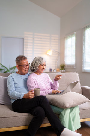 Cheerful older Asian couple is using a laptop in their cozy home. They are enjoying retirement together while browsing the internet and staying connected online.の写真素材
