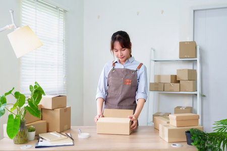 An Asian young woman carefully packing merchandise into boxes for delivery. She is working from home fulfilling orders for her online business to customers.の写真素材