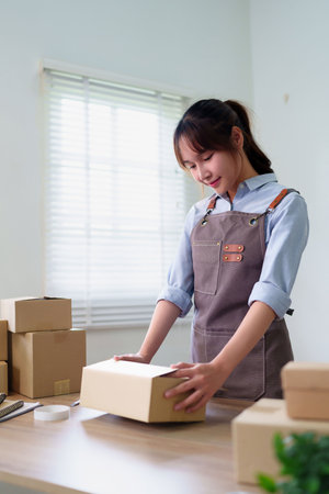 A young Asian woman carefully packing a cardboard box at her home for delivery. She's a small business owner preparing an order to ship for her online store.の写真素材