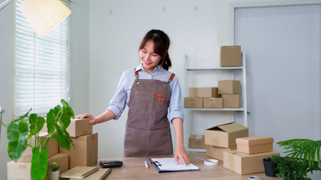 A smiling Asian young woman in an apron prepares packages for shipping working from her home office providing top quality service to her online shop customers.の写真素材