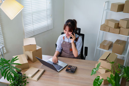 Asian woman entrepreneur manages her small business from home using a laptop and phone to handle orders and shipping. She ensures smooth ecommerce and delivery.の写真素材
