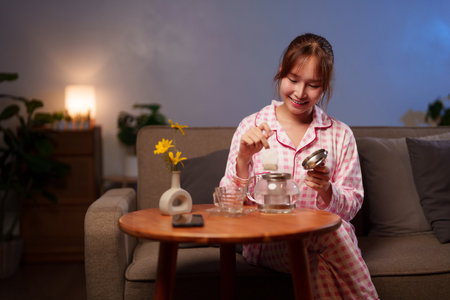 An adult Asian woman in pajamas prepares tea, showcasing a cozy relaxation ritual and promoting longevity through healthy lifestyle choices with a tea infusion at home.の写真素材