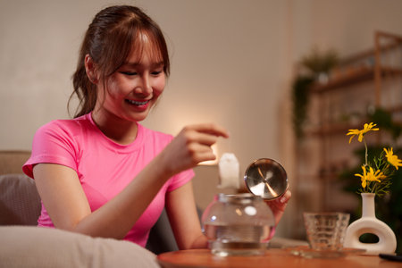 A young Asian woman prepares healthy herbal tea infusion in a teapot for her longevity lifestyle routine, enhancing calm and well being near a vase with flowers.の写真素材