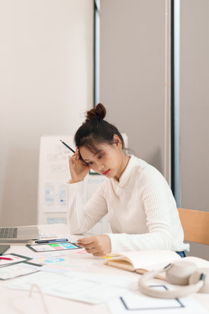 Asian adult woman UI designer is working hard on her design project in the office, creating application design with laptop and paper wireframes for a mobile appの写真素材