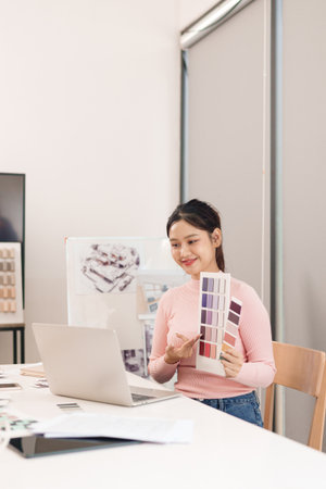 Young adult Asian woman UI designer sits at her desk with laptop and color swatches. She smiles as she selects colors for her next mobile app project.の写真素材