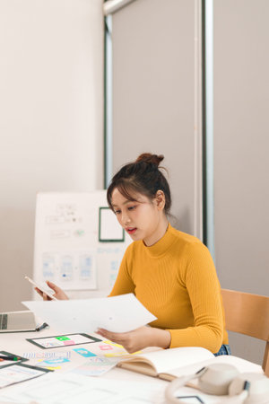 Adult Asian woman UI UX designer creating mobile app interface design. She uses mobile phone and...