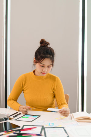 An adult Asian woman working as a UI UX designer is developing a mobile app interface and focusing...