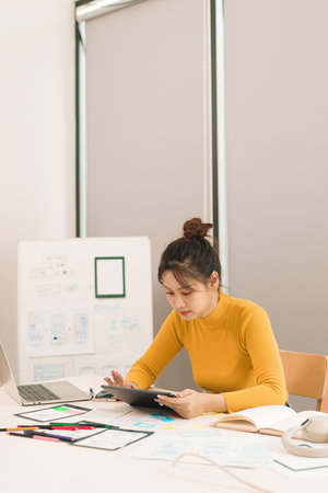 Adult Asian woman user interface designer working with a tablet to create innovative mobile app interfaces for a smartphone project focusing on creativity and website usabilityの写真素材