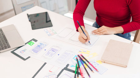 Adult Asian woman designs user interface for mobile application on the desk. She uses a laptop,...