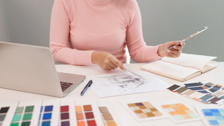 Asian woman works on UI UX design in a bright workspace. She is selecting colors for an interior design project while using a laptop and color palette.の写真素材