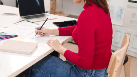 An Asian woman in a red sweater is sketching a mobile app wireframe for user interface design on a...