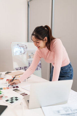A young Asian woman is working as UI UX designer in her creative studio. She uses a laptop and color...