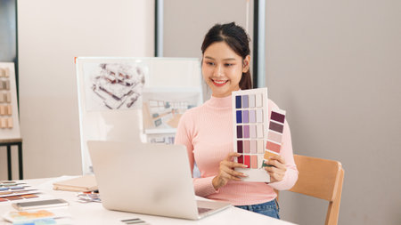 Asian young adult woman interior designer choosing color palette at desk with laptop during online meeting. Working on an interior design project at home.の写真素材