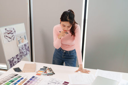 A young Asian woman is working as a UI designer reviewing floor plans color swatches and sketching...