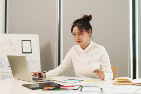 Young adult Asian woman UI UX designer is working on a laptop for mobile application and website interface project. Designing user experience in the office.の写真素材
