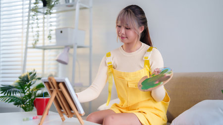Capture the essence of creativity as a young Asian woman skillfully paints on canvas. She is holding palette with colors and wears yellow dress to showcase her style.の写真素材