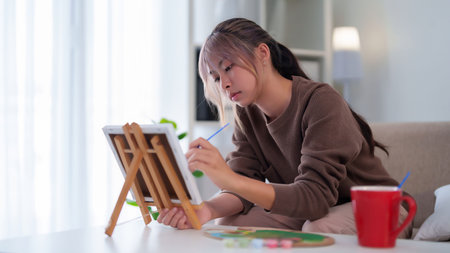 Captivating image of a young Asian woman deeply engrossed in her artwork She uses a paintbrush to...