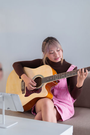 Adult Asian woman plays acoustic guitar sitting on sofa learning music chords online course happy...